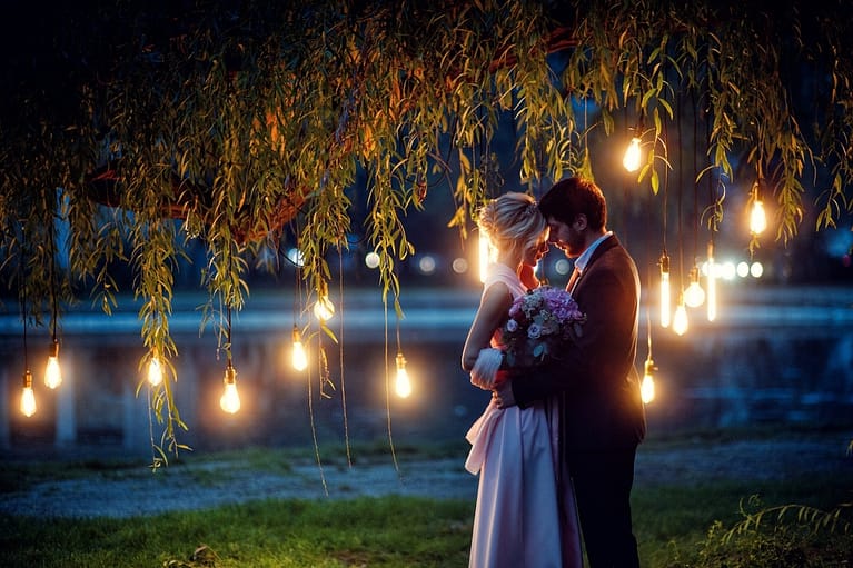 Fairy night portrait of wedding couple with lights of edison lamps by blue lake under willow tree